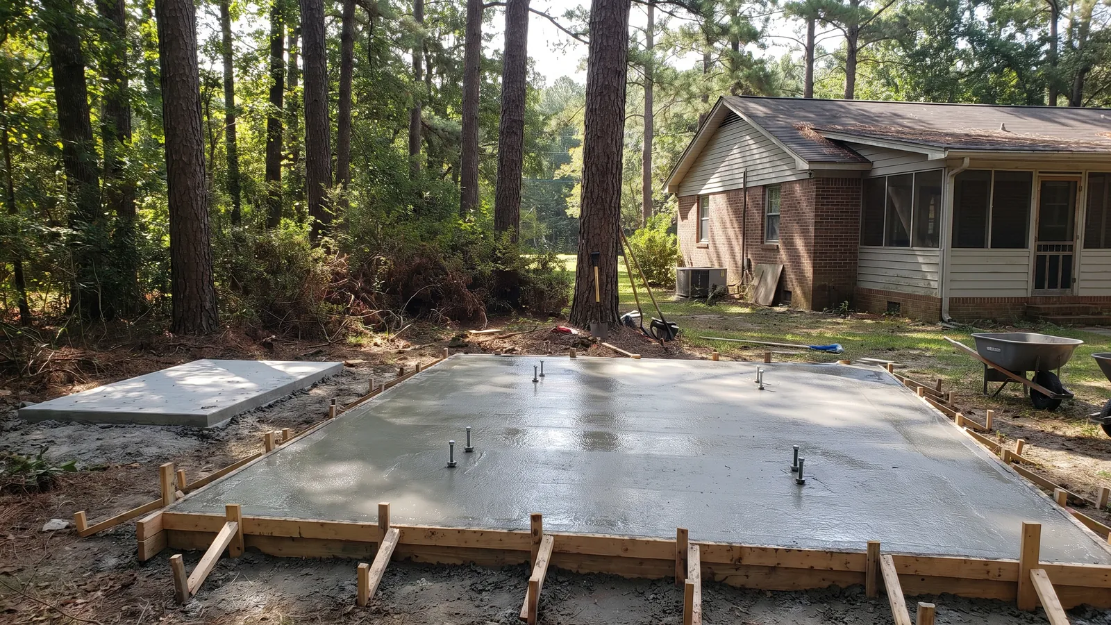 Freshly poured reinforced concrete pad with anchor bolts beside a standby generator pad in a pine-shaded South Carolina backyard